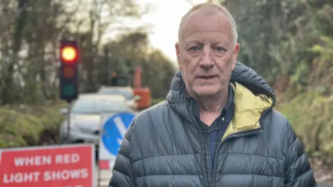 Frankie is standing on the Glenshesk Road, he is wearing a dark waterproof coat which has a yellow lining on the inside of the hood. Frankie has grey hair. He is standing in front of temporary traffic controls. The light is red and there is a sign which reads "when red light shows wait here".