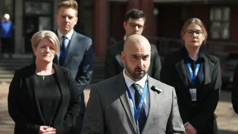 Supt Simon Vickers with a shaved head and dark beard wearing a light grey suit and dark blue tie reads a statement on the steps of the court surrounded by other police officers