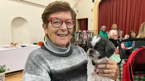 BBC A woman in red framed glasses holding a small grey dog on her lap. She is sitting in a village hall