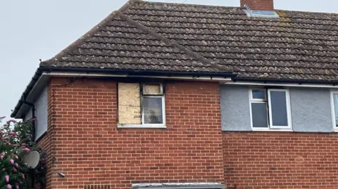 A brown brick house with a board covering one of the windows. Round both of the window flames you can see black smoke damage.