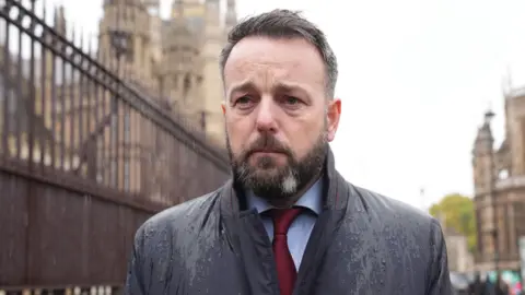 PA Media A man with black and grey hair, and beard, is standing outside looking to the left of the camera. He is wearing a coat, wet from rain, a maroon tie and light blue shirt. A fence can be seen behind him as can an old tall building. 