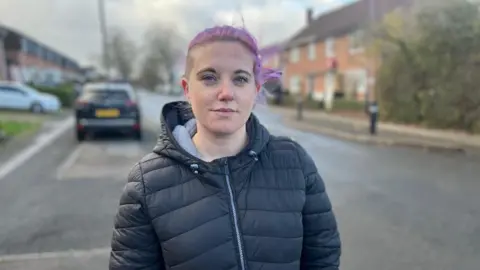 BBC Charlie Ward is wearing a black jacket. She is standing by a road, with two cars behind her. 