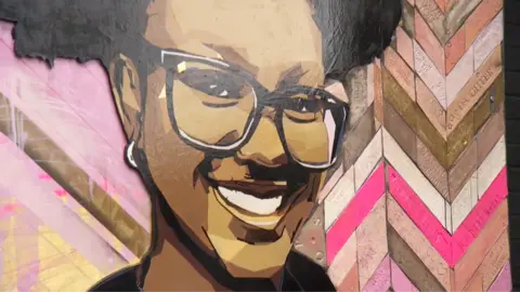 The mural of Elianne smiling and wearing glasses. To the side of her portrait are ceramic tiles, with inscriptions from her family and friends, including 'we love you Elianne'.  