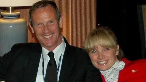 Louise Woollam A younger Richard and Louise Woollam are sitting on a sofa. He has short grey-brown hair and a pilot's uniform. She has blonde hair in a fringe and wears the red uniform of an airline's cabin crew.