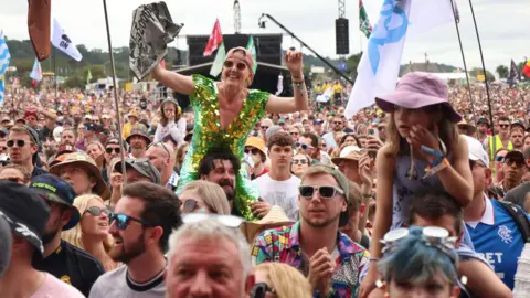 A large crowd of people watch a performer on Glastonbury's Pyramid Stage, they are wearing a variety of colourful clothing and sunglasses