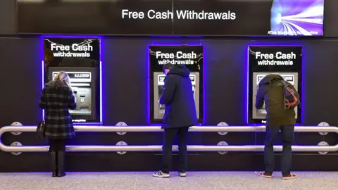 Three people (one woman, one man and one person out of view) withdraw cash from free-to-use cash machines