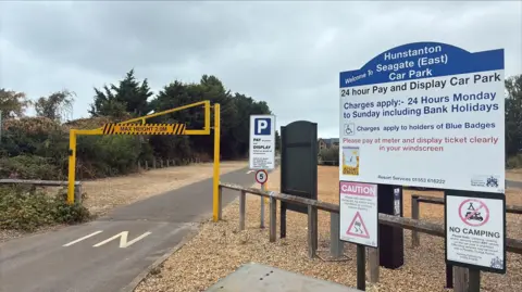 Robby West/BBC Photograph shows the entrance to the Seagate (East) Car Park in Hunstanton, with a blue and white information sign to the right. Also in view are signs warning the site is a "NO CAMPING" zone and there is a yellow height restriction frame visible at the entrance.