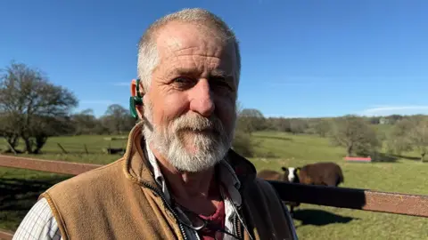 Farmer Mark Firby is standing by the rung of a metal gate with a field behind him. In the grassy field are badger face Welsh mountain sheep and a handful of small trees. Firby has short, white-grey hair, a moustache and beard. He's wearing a cream checked shirt and a beige, zip-up, sleeveless vest. On his right ear is a green earpiece.