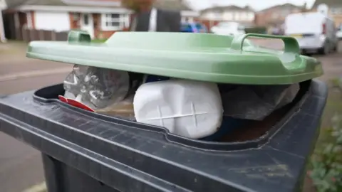 BBC A black bin with a green lid so full with plastic recycling waste that the lid will not close