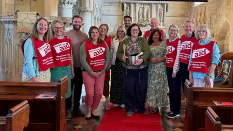 Phil Juggins A group of adults wearing red tabards with the words 'Christian Aid' written on them, while standing in a church. The woman in the middle is holding a cooking pot and a wooden spoon.