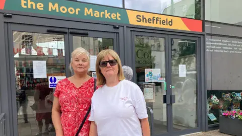 Naj Modak/BBC Kay and Sheila have blonde hair. Kay has a patterned dress and Sheila has a white top and sunglasses. They are stood in front of a glass building with a sign that reads, The Moor Market, Sheffield.