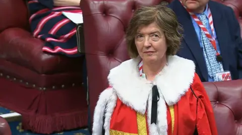 PA Media Sue Gray wears a red and white ceremonial robe with gold trim in the House of Lords surrounded by seated individuals on red leather tufted benches. The room features a blue carpet and ornate decorations, suggesting an official or governmental setting.