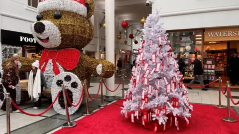 A white Christmas tree covered in red labels stands on a red carpet in front of a Waterstones store. A large teddy bear wearing a Santa hat and a red bowtie sits next to it