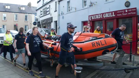 RNLI A group of eight people are pulling a bright orange life boat through a narrow street. the boat is mounted on a trailer. the boat says RNLI across the front.