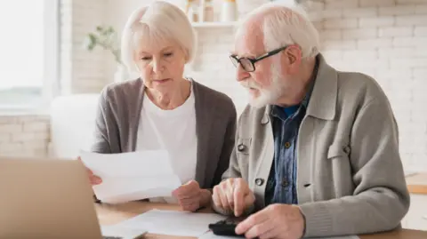 Getty Images Older man and woman sit at a table in a kitchen with paperwork and a laptop in front of them