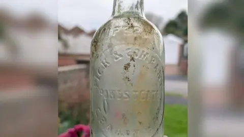 David Martin/Grantham RiverCare A clear thin-necked bottle with mud on it being held up. An inscription on the front reads "Imperial, Pick & Company, Westgate, Grantham."