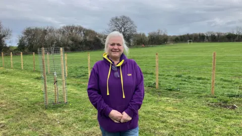 A woman in a purple and yellow hoodie, with glasses on the collar, stand centrally looking into the camera. Behind her is a green field of grass.
