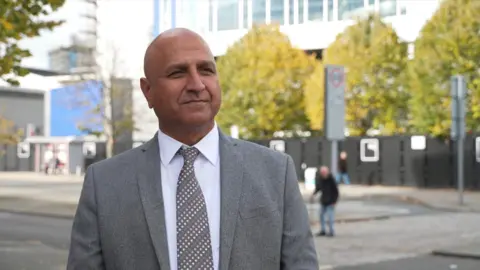 Councillor Naeem Akhtar stands in Coventry city centre. He is wearing a grey suit jacket, a grey, pink and white striped tie and a white shirt. 