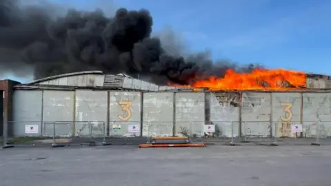 Roger Leggard Orange flames and tall plumes of black smoke billowing out from the top of a hangar building.