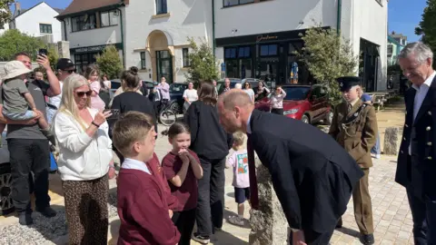 Prince William speaking to schoolhouse children who are stood successful beforehand of a crowd. There's a pistillate stood down them taking a photograph of them each talking.