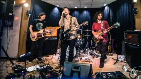 Joe Bamford A band play on a stage surrounded by sound equipment. Three band members are standing and each holding a guitar. The background is a black curtain with lights and behind the guitarists, there is a drummer sitting at a drum kit.