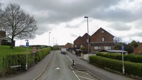 A one-way street, which has blue signs with white arrows indicating the direction of travel, and a speed bump along with other traffic calming measures. There are hedges either side.