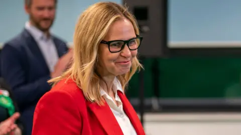 PA Media Helen Godwin on the night of her election. She wears a red suit, has shoulder length blond hair and glasses. 