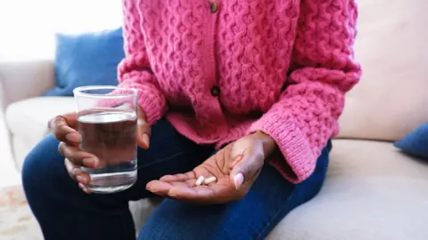 Getty Images Woman wearing bright pink cardigan and dark blue jeans hold two pills in one hand and a glass of water in the other 