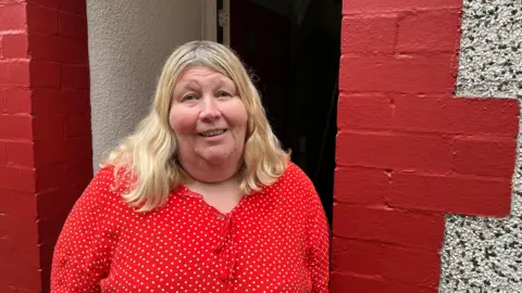 A woman looks at the camera with a wry smile. she is standing in the doorway of a red brick home. she is wearing a red top with dots and has shoulder length blonde hair