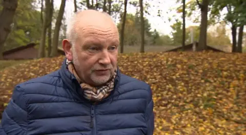 A man wearing a blue coat and brown scarf looks away from the camera. Autumn leaves and trees are in the background.