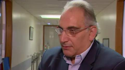 BBC Professor Derek Manas wearing glasses, a dark blue blazer and a purple shirt. He has grey hair, swept back. He is standing in a hospital corridor