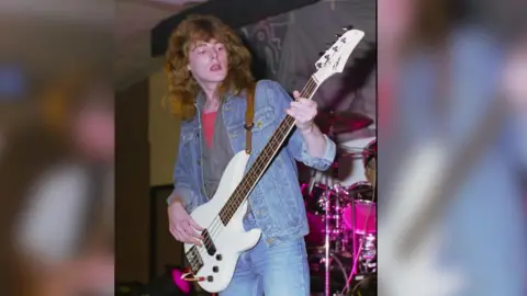 James Lewis His father is pictured as a young man playing a white guitar on a stage with a drummer behind him. He is wearing jeans and a denim jacket and a top underneath. He has long reddish/brown hair.