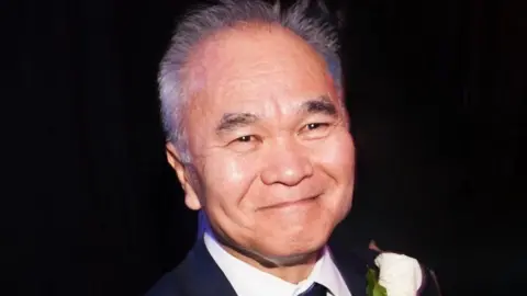 Close up of Danny Boon with grey hair and wearing a black suit, white shirt and black tie with a white rose boutonniere. He is smiling.
