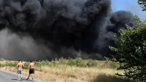 Reuters Two men walk past a big grey cloud of smoke, the site of Arnolds Field in Launders Lane, Rainham