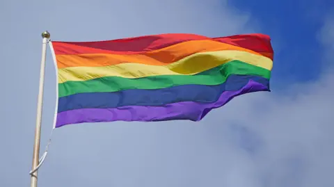 A rainbow-coloured Pride flag flying against a blue sky with light clouds.