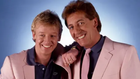 Les Dennis and Dustin Gee standing together and smiling in matching pink suits