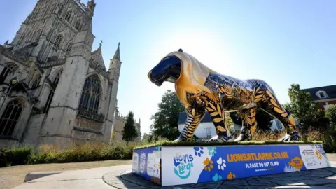 A lion sculpture painted in black and gold is stood in front of Gloucester cathedral. The sun is shining and the sky is blue.