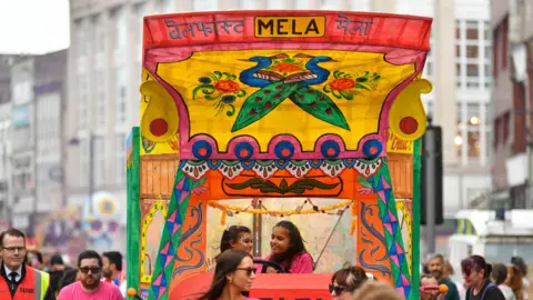 Pacemaker Two young girls on a Mela float, which has loads of colour and has peacocks on the front of the cover 