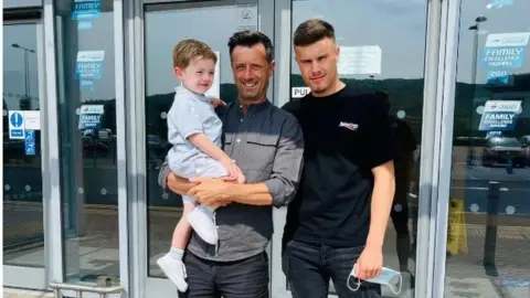 Jason Bowen Jason Bowen, wearing a grey shirt and black trousers, holds a young child. To his right is a young man wearing a black t-shirt and black jeans