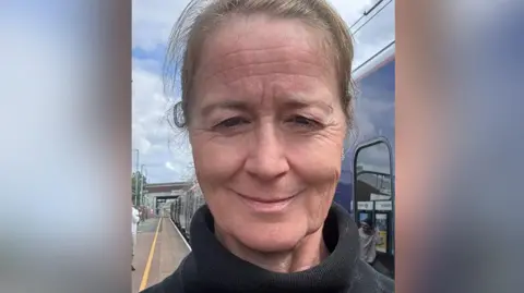 Sarah Nelson Sarah Nelson selfie on a railway station platform after having to come off a train. She has light brown hair worn back in a ponytail and wears a black polo neck jumper. She is smiling. 