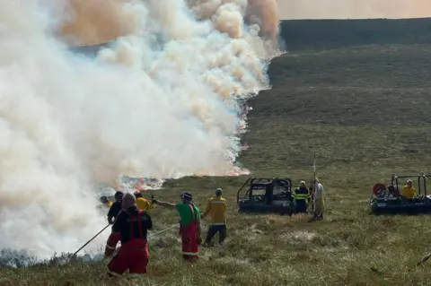 RSPB Scotland Firefighters and volunteers work to contain a large moorland wildfire as a long line of flames advances across a hillside. Thick plumes of white and brown smoke rise into the sky while crews use hoses and equipment vehicles positioned on the grassland below.