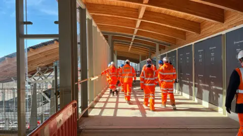 Tom Jackson/BBC Construction workers in orange hi-vis overalls and hard hats are walking on a bridge at a railway station which goes above the railway lines.