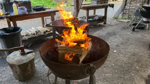 Fire is visible in a bowl which is above a larger bowl that is outside. Benches have objects on in the background and a bin is nearby.
