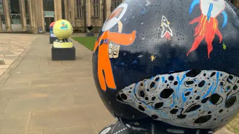 A dark coloured globe depicts images by children of space travel. It is on a plinth outdoors with other globes in the background