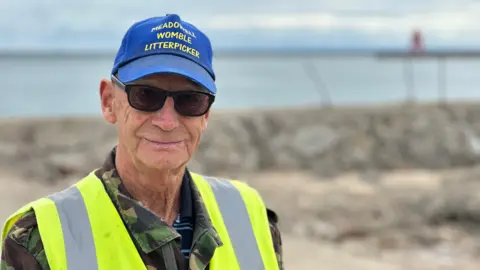 Dave Shaw is standing facing away from the beach and water, which are out of focus behind him. He is wearing a blue baseball hat with yellow lettering saying he's a "Womble litter picker" and is wearing a high-vis jacket over a camouflage jacket. He is wearing sunglasses and is half smiling.