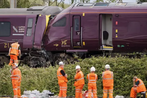 Two trains are seen crashed together with people wearing orange protective clothing standing alongside.