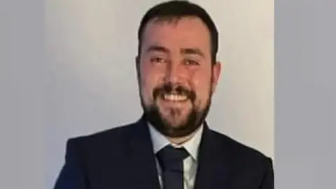 Police Scotland Smiling man with a beard wearing a dark suit and tie against a plain light-coloured background.
