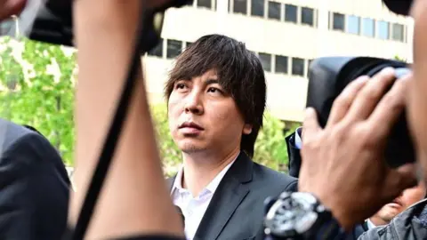 Getty Images Ippei Mizuhara seen outside a California courthouse. Photographers surround him and he's wearing a white button-down shirt and black suit jacket. 