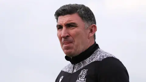 Getty Images Kevin Maher standing in front of a grey sky while licking his lips and looking focused. He has dark, short hair and is wearing a black and white Southend United tracksuit jacket.
