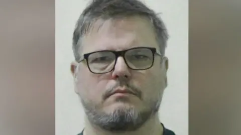 Mugshot of Dean Juric. He has scruffy dark and grey hair and a black and grey beard and is wearing black-framed glasses